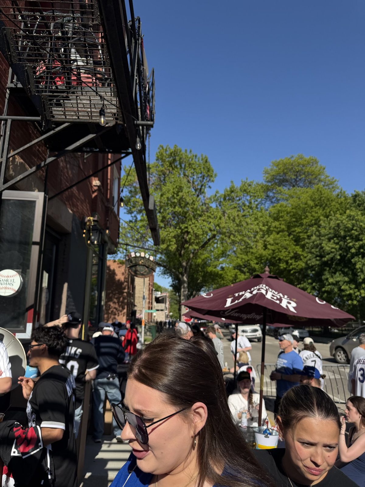 The outdoor beer garden at Cork & Kerry at the Park near Rate Field on a sunny pre-game evening, with Sox fans seated under umbrellas and a Bridgeport streetscape in the background